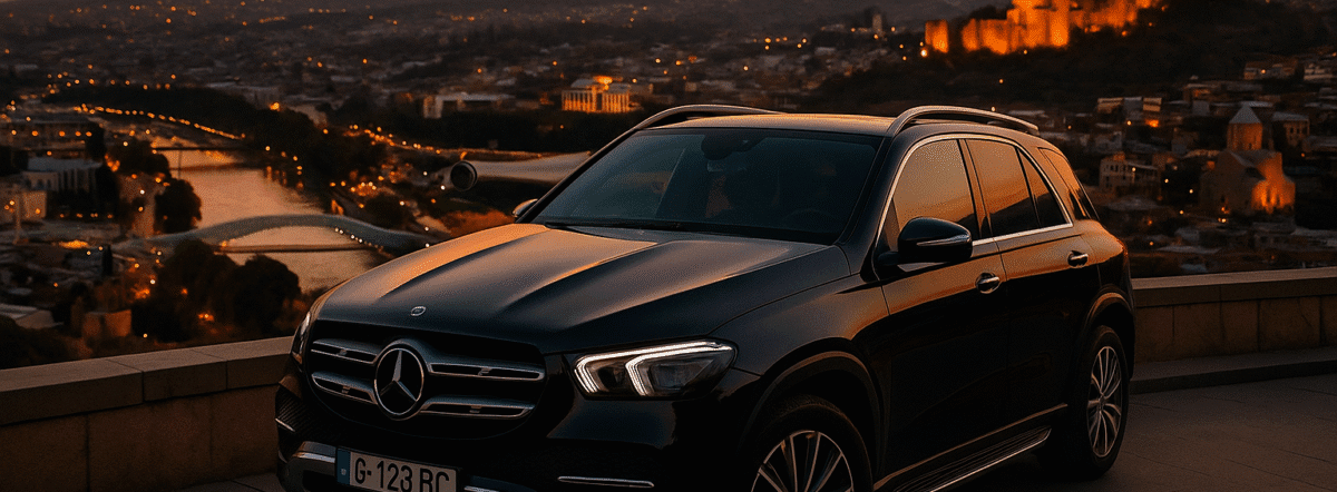 Black Mercedes-Benz GLE SUV parked with Tbilisi city skyline and Narikala Fortress in the background at sunset.